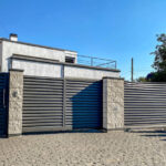 A gray private house of two floors hidden behind a fence with automatic sliding brown gates and an area near the house against a blue sky.